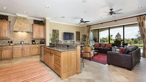 A spacious kitchen with wooden cabinets, granite countertops, and a central island. There is a dining area with a sofa and a large window offering a view of the outdoors.