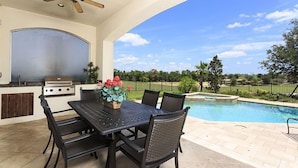 An outdoor patio with a table and chairs, a grill, and a view of a swimming pool.