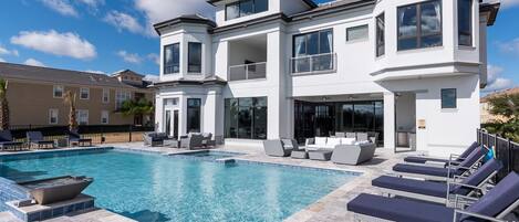 A modern two-story house with a swimming pool, outdoor seating, and a large window overlooking the pool.