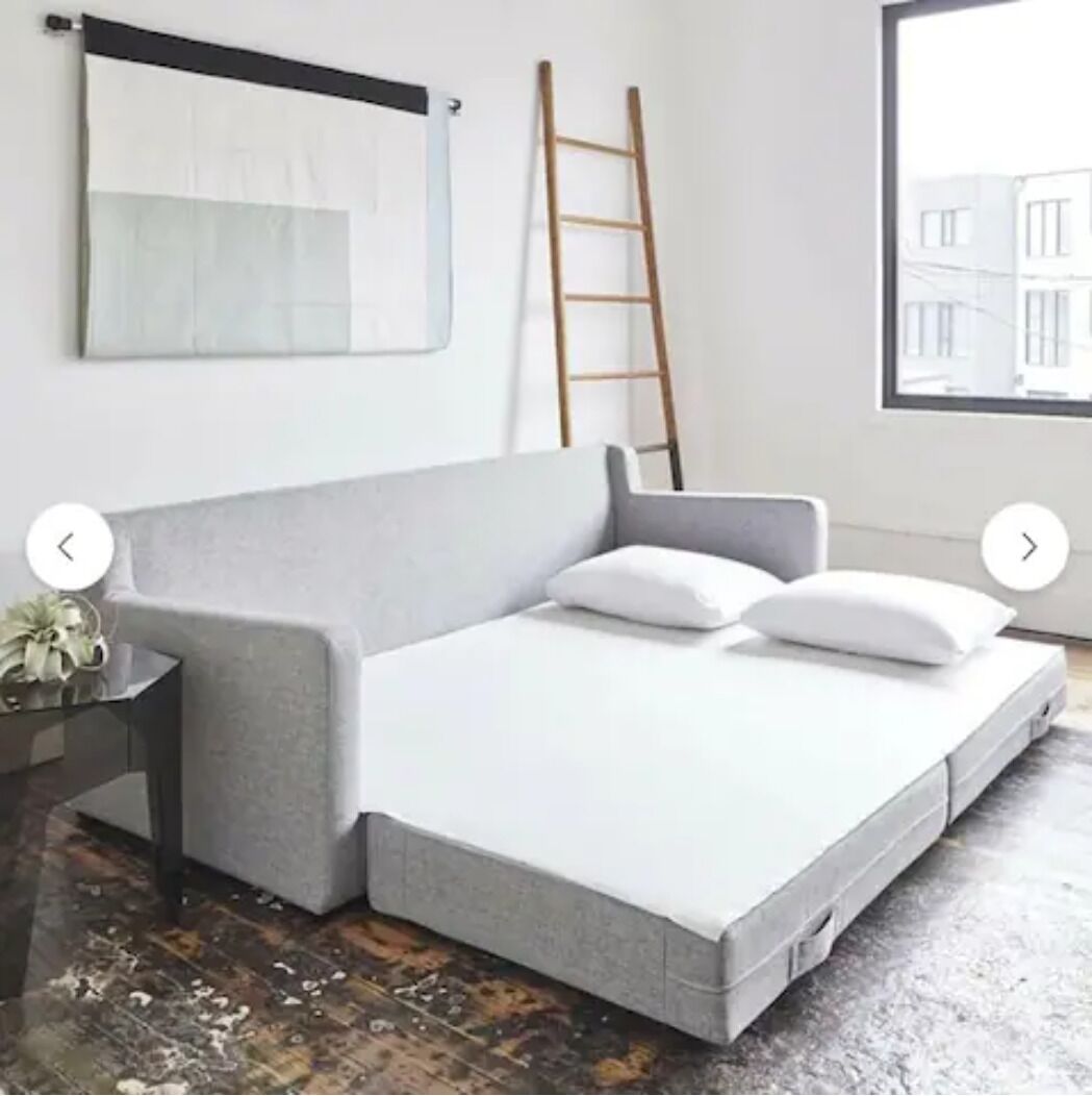 A modern bedroom with a grey sofa bed, a wooden ladder, and a large window with a city view.