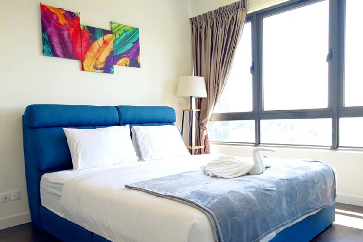 A hotel room with a large bed, a window with curtains, and two colorful wall art pieces.