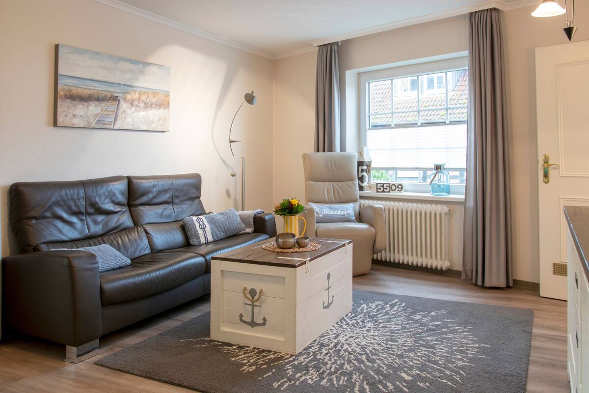 A living room with a leather sofa, a coffee table with an anchor design, a painting on the wall, a window with curtains, and a radiator.