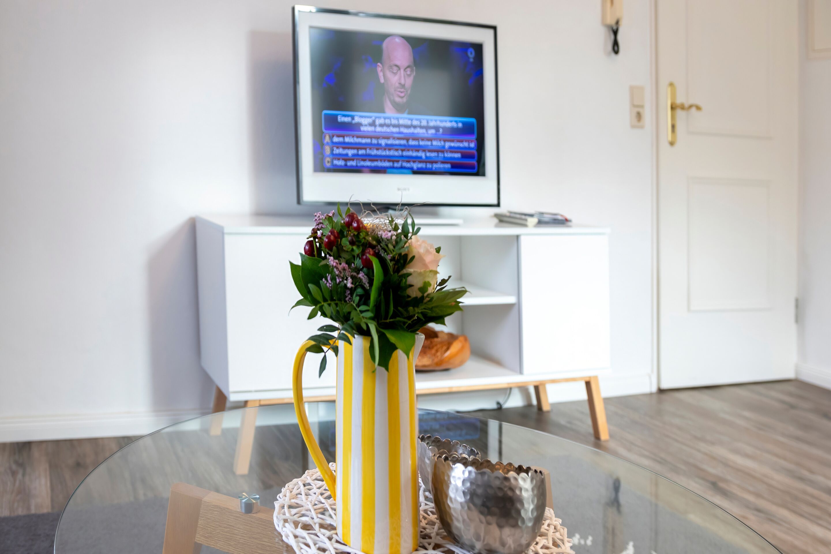 A room with a glass coffee table, a yellow striped vase with flowers, a metallic bowl, and a white entertainment unit with a television displaying a news channel.