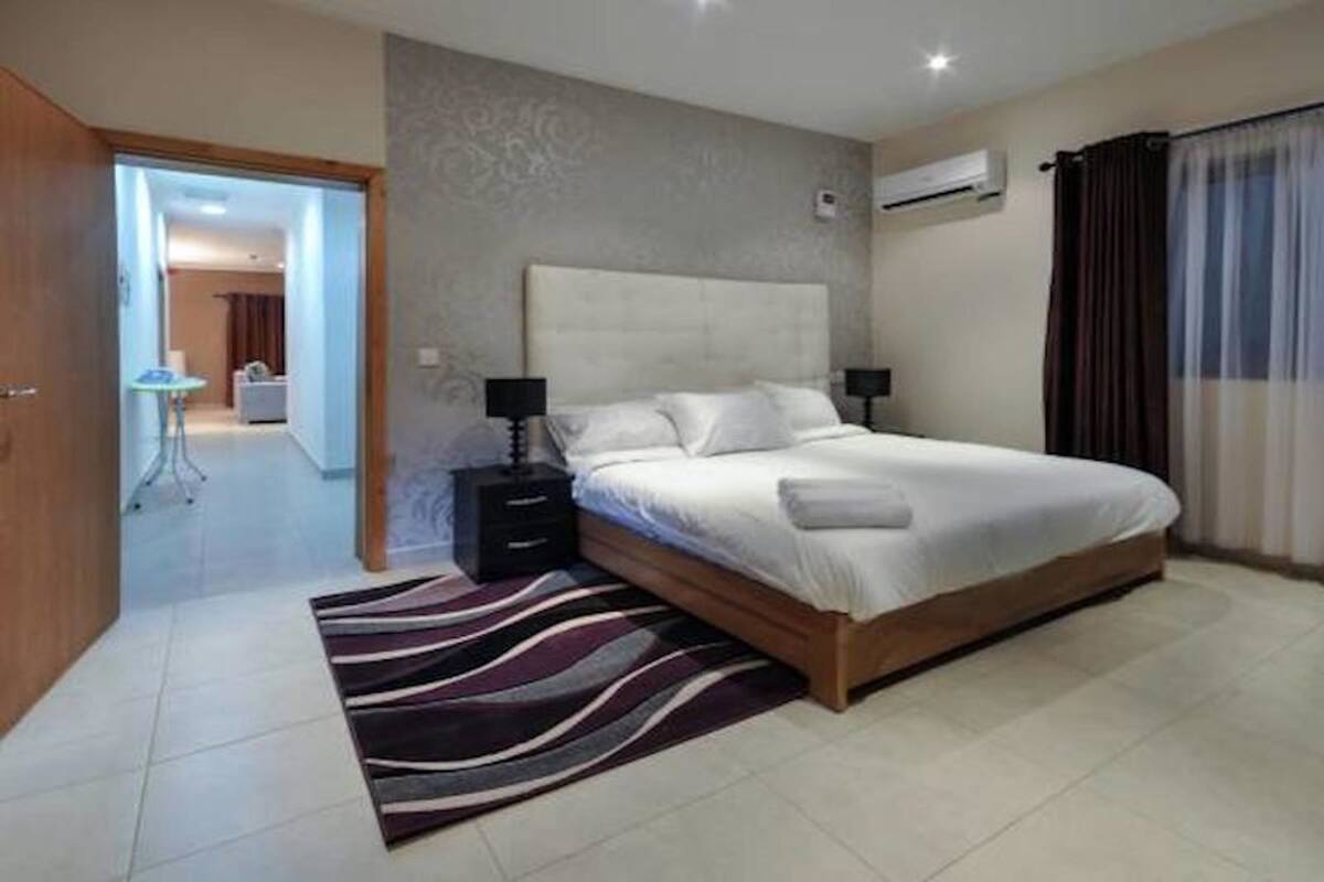 A bedroom with a large bed, two bedside tables, a wall-mounted air conditioner, and a view of the hallway.