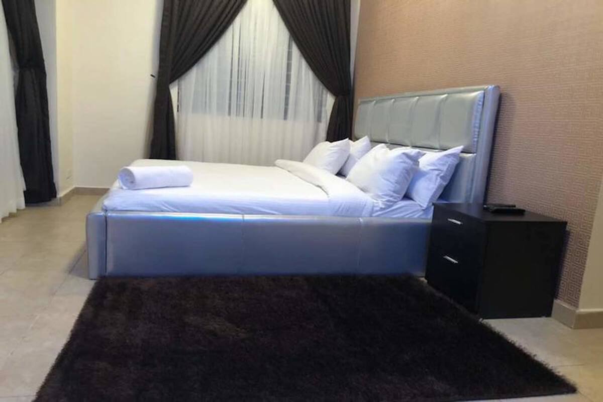 A neatly made bed with white linens, a dark brown rug, and a black nightstand with a remote control.