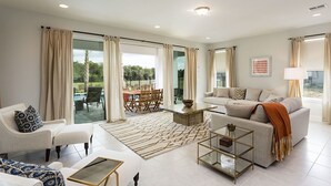 A spacious living room with a sofa, armchair, and coffee table, large windows with curtains, and a view of the outdoors.
