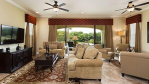 Un salón espacioso con un televisor de pantalla plana grande, dos ventiladores de techo y vistas al exterior con mucha vegetación.