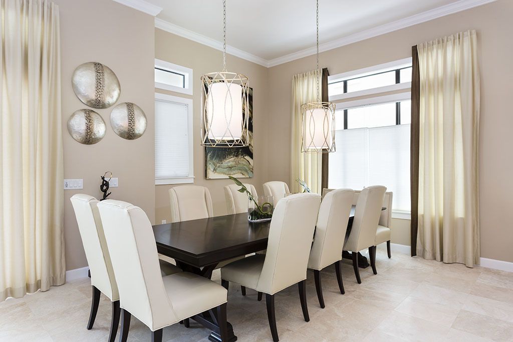 A dining room with a dark wooden table surrounded by six chairs, large windows with beige curtains, and two hanging light fixtures.