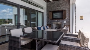 A modern outdoor patio with a glass enclosure, wicker furniture, a flat-screen TV, and a stone fireplace.