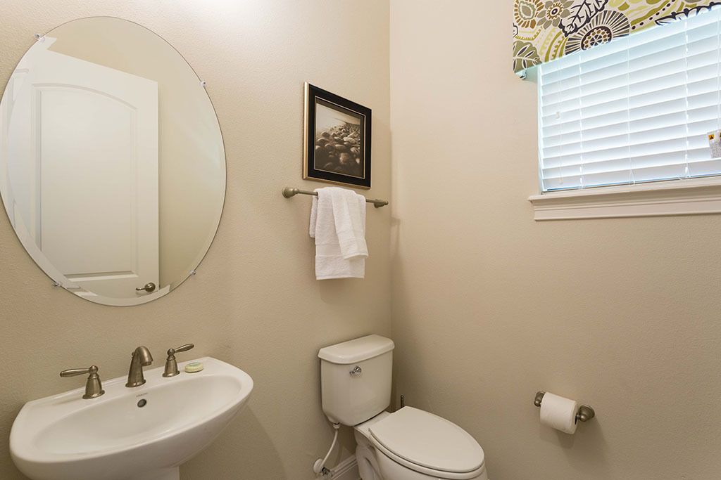 Een badkamer met een ronde spiegel, een wastafel, een toilet, een afbeelding in een lijst en een raam met jaloezieën.