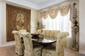 A dining room with a round table, floral upholstered chairs, a large tapestry, and a window with draped curtains.