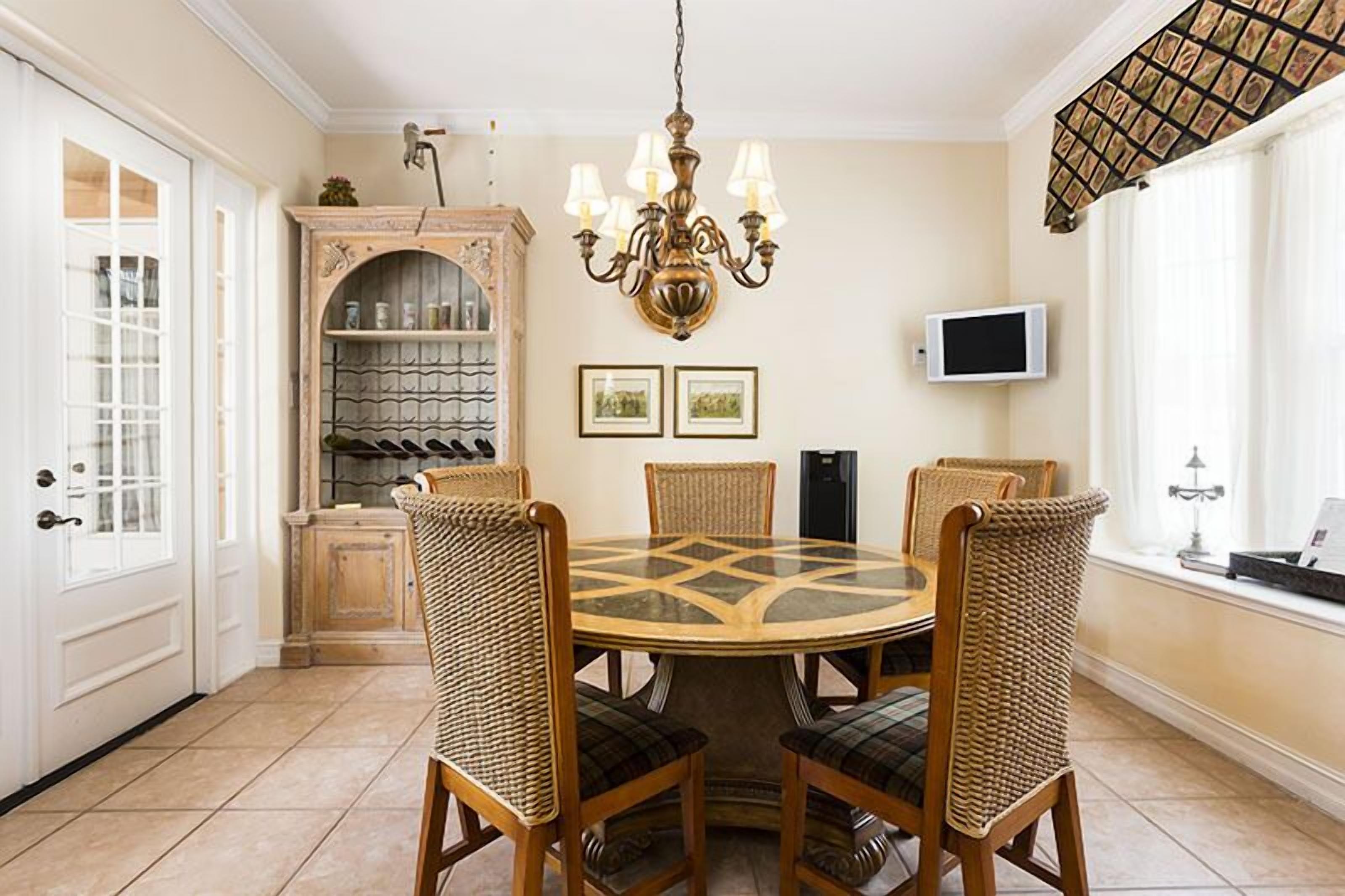 Un comedor con una mesa redonda, cuatro sillas de mimbre, una araña, una vinoteca y un televisor en la pared.