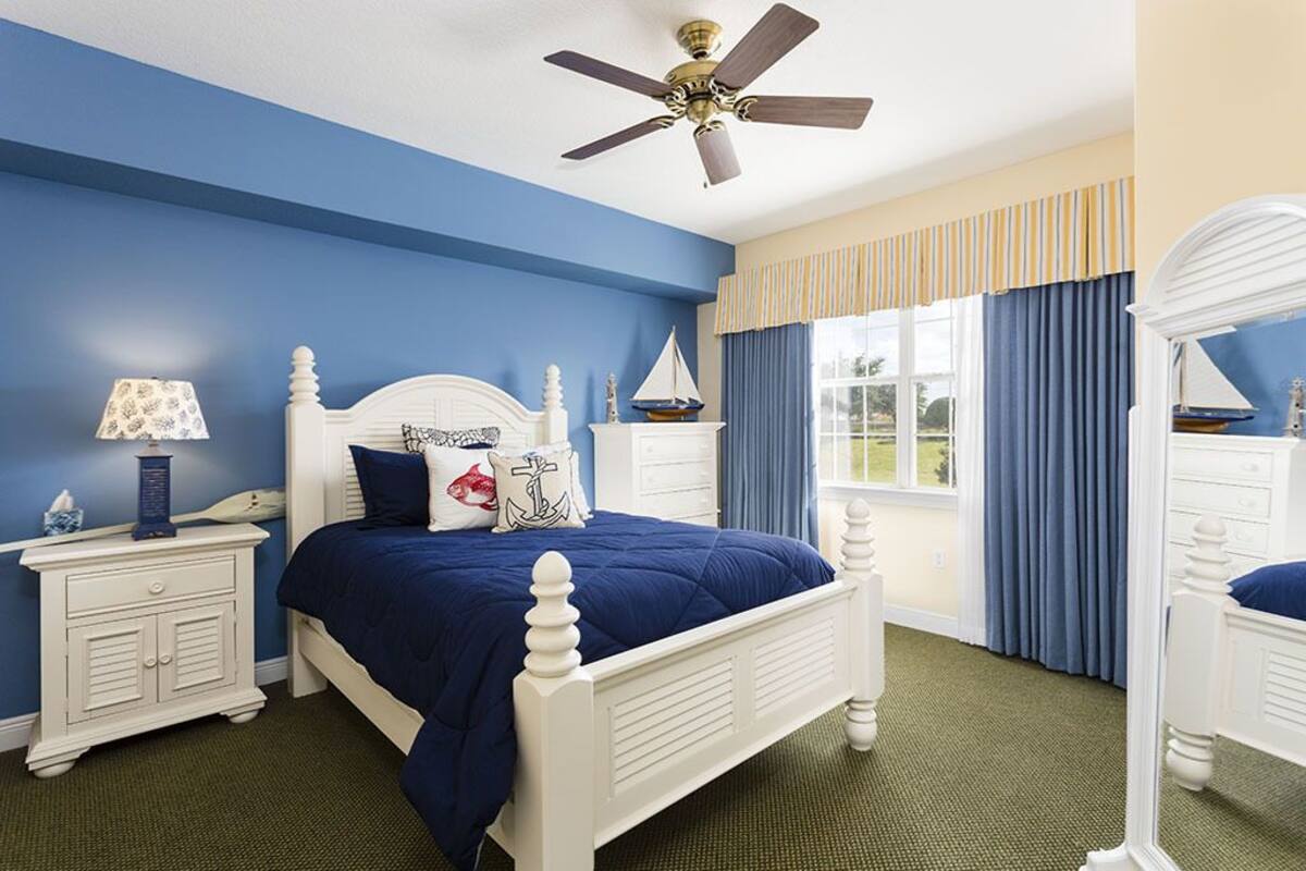 Une chambre avec un lit blanc, une literie bleu marine, un ventilateur de plafond, une table de chevet avec une lampe et une fenêtre avec des rideaux.
