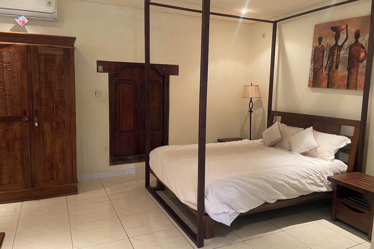 Uma cama com dossel, roupa de cama branca, um guarda-roupa de madeira, um candeeiro de chão e um quadro na parede.