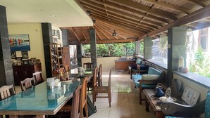 A spacious dining area with a glass-top table, wooden chairs, and a seating area with cushioned sofas and a small bar. - Graha Madesimon (Batubulan)