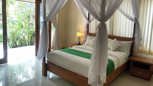 A room with a bed, a nightstand, a lamp, and a view of greenery outside. - Graha Madesimon (Batubulan)