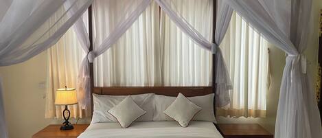 A bed with white bedding, a green throw, and a wooden headboard. There is a nightstand with a lamp and a wooden table beside the bed.