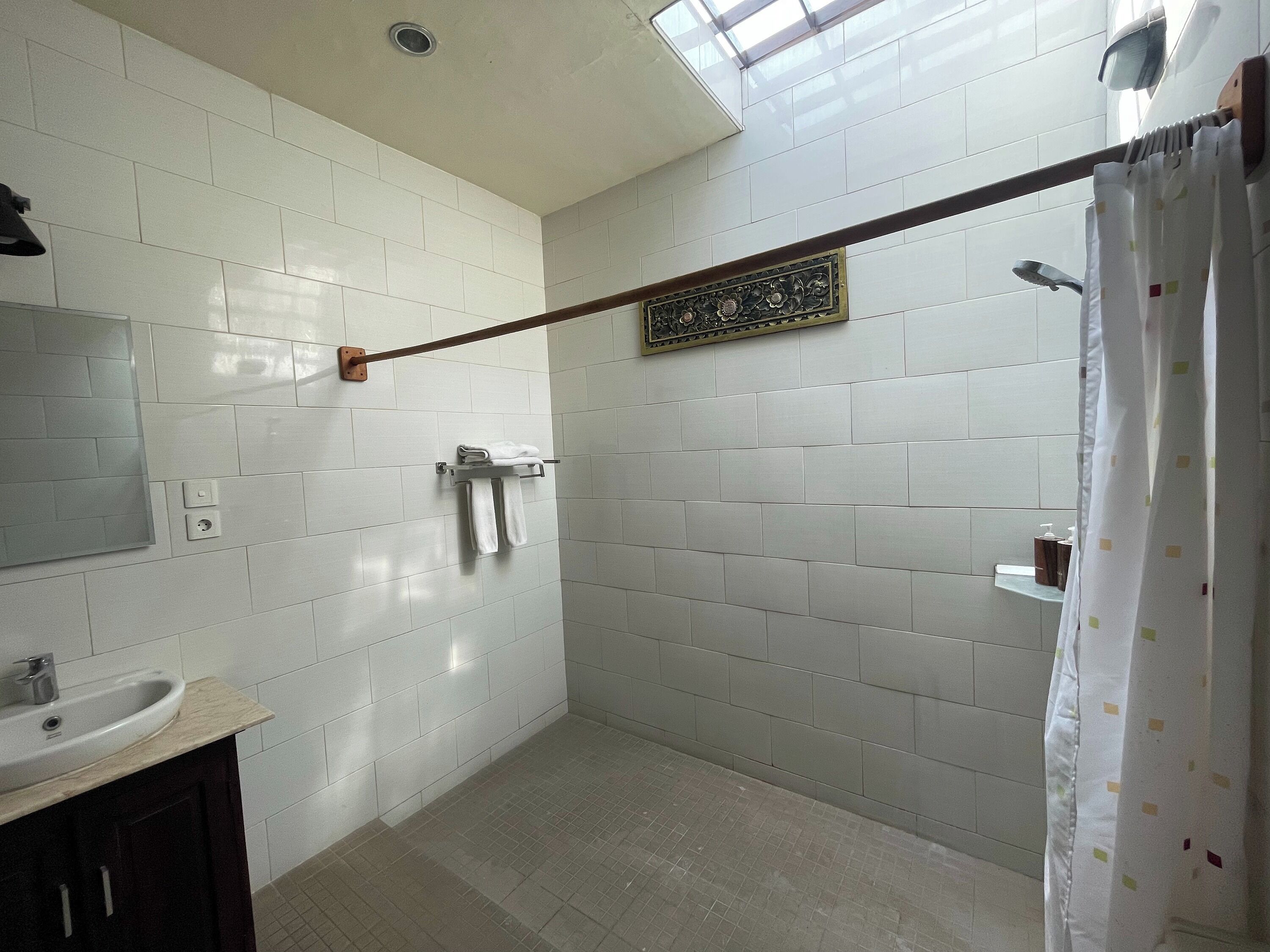 Un bagno con pareti piastrellate di bianco, un portasciugamani in legno, uno specchio, un lavandino e una tenda da doccia.