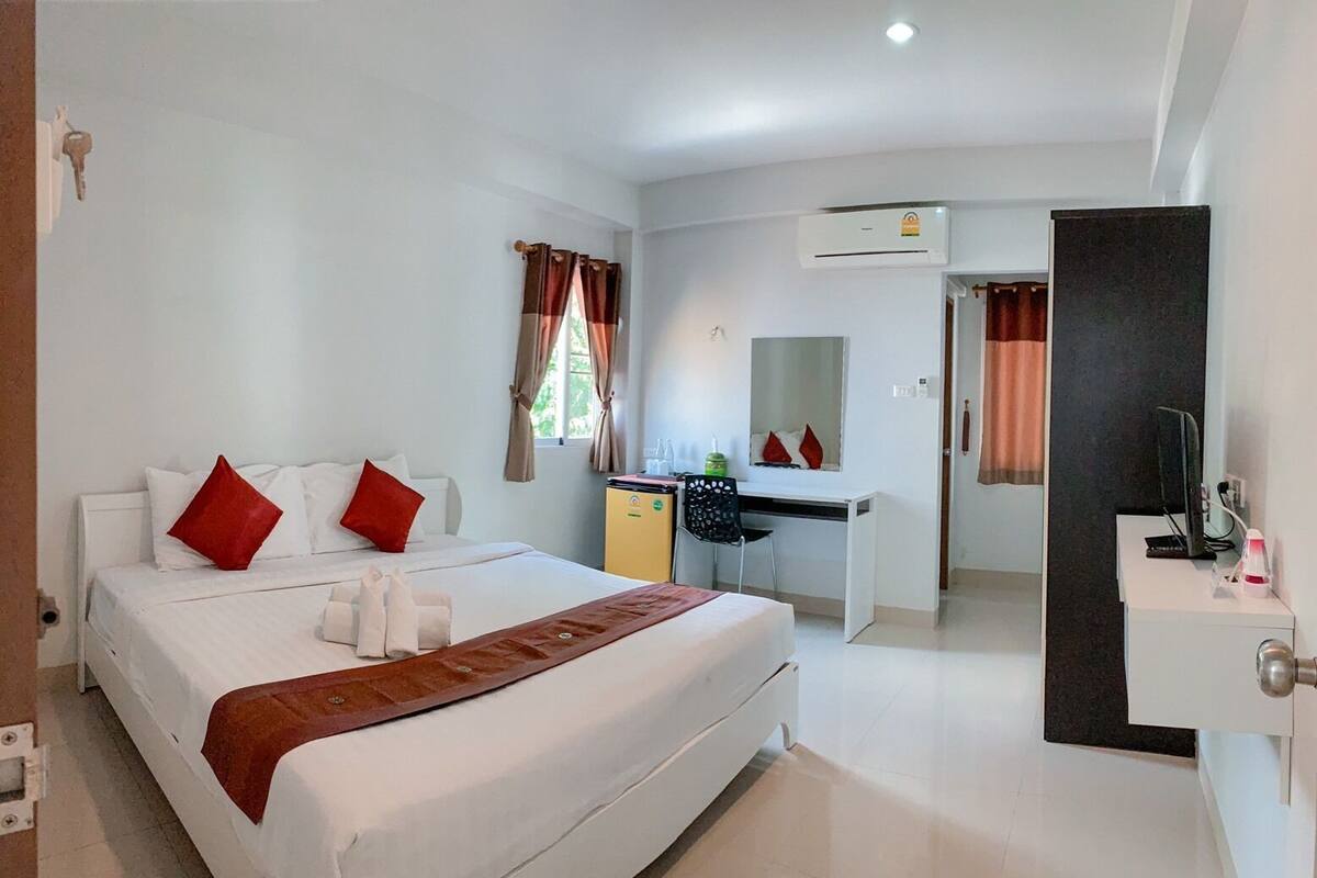 A hotel room with a bed, a desk, a chair, a TV, and an air conditioning unit.
