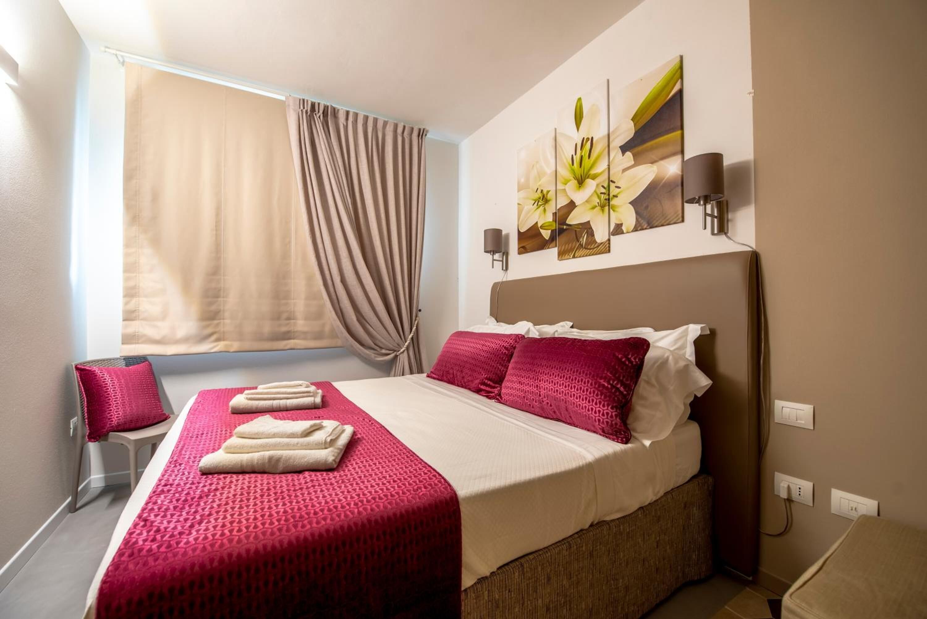 Una camera d'albergo con un letto, una finestra con tende, una lampada a parete e un quadro con fiori.