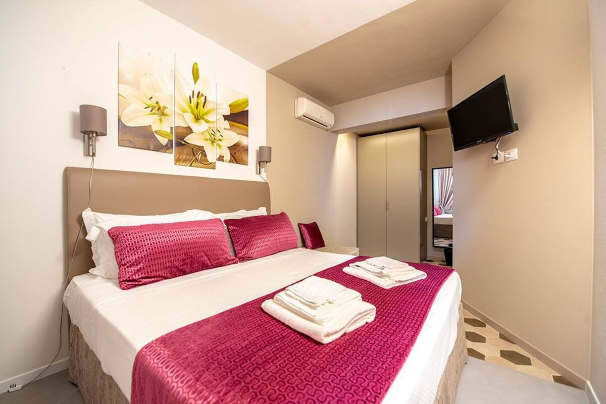 A hotel room with a bed, a flat-screen TV mounted on the wall, and a painting of flowers above the bed.