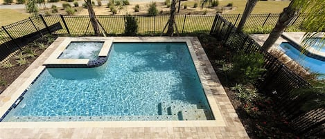 A rectangular swimming pool with a hot tub, surrounded by a brick deck and a black metal fence.