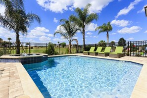 Et rektangulært swimmingpool med krystalklart, blåt vand, omgivet af en flisbelagt terrasse og et hegn i sort metal. Tre grønne liggestole står langs poolkanten.