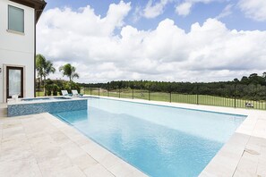 Une piscine rectangulaire avec une terrasse carrelée, un bâtiment blanc doté d’une fenêtre et d’une porte, et une clôture délimitant une zone herbeuse avec des arbres.