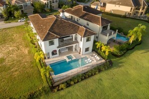 Una casa grande de dos pisos con piscina, rodeada de un césped cuidado y palmeras.