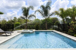 Una piscina rectangular con jacuzzi, rodeada por una valla negra, palmeras y un cielo despejado.