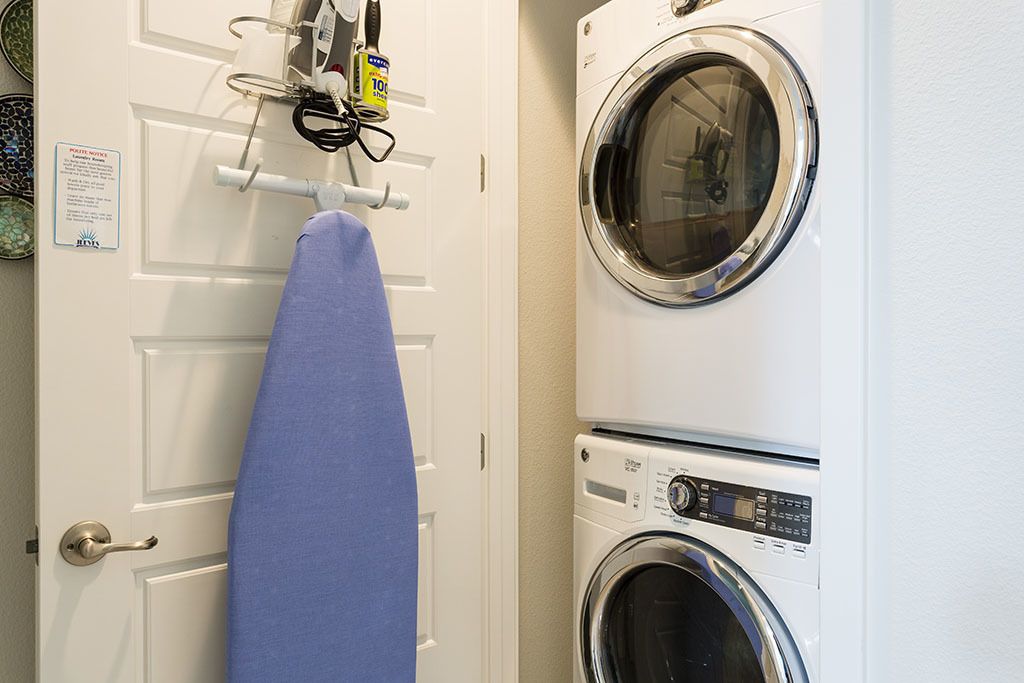 A laundry area with a washing machine and dryer, a blue ironing board, and a notice on the door.