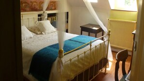 A bedroom with a four-poster bed, a nightstand, a chair, and a window with a view of greenery. - La Petite Vigne (Amboise)