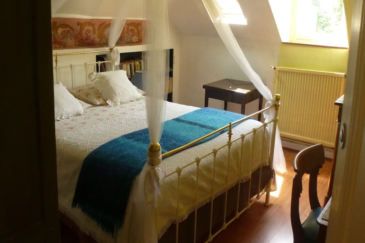 A bedroom with a four-poster bed, a nightstand, a chair, and a window with a view of greenery.