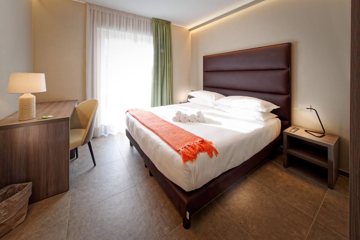 Et hotelværelse med seng, sengebord, lampe, stol, skrivebord og gardiner.