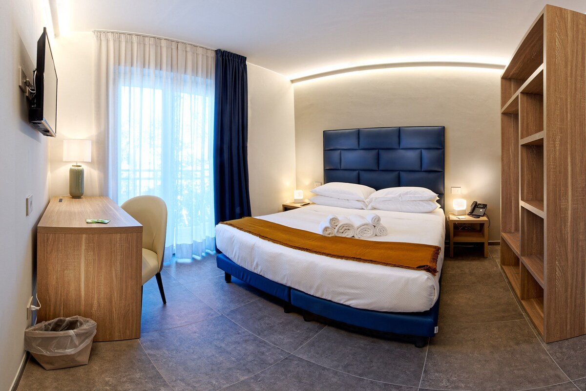 A hotel room with a bed, bedside tables, a desk, a chair, a TV, and a wardrobe.