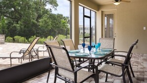 A covered patio with a table set for four, surrounded by chairs, and a view of trees and a fence in the background. - Je62429 - Reunion Resort - 8 Bed 8 Baths Villa (Kissimmee)