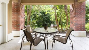 Un patio cubierto con una mesa redonda de vidrio y cuatro sillas, rodeado de pilares de ladrillo y abundante vegetación.