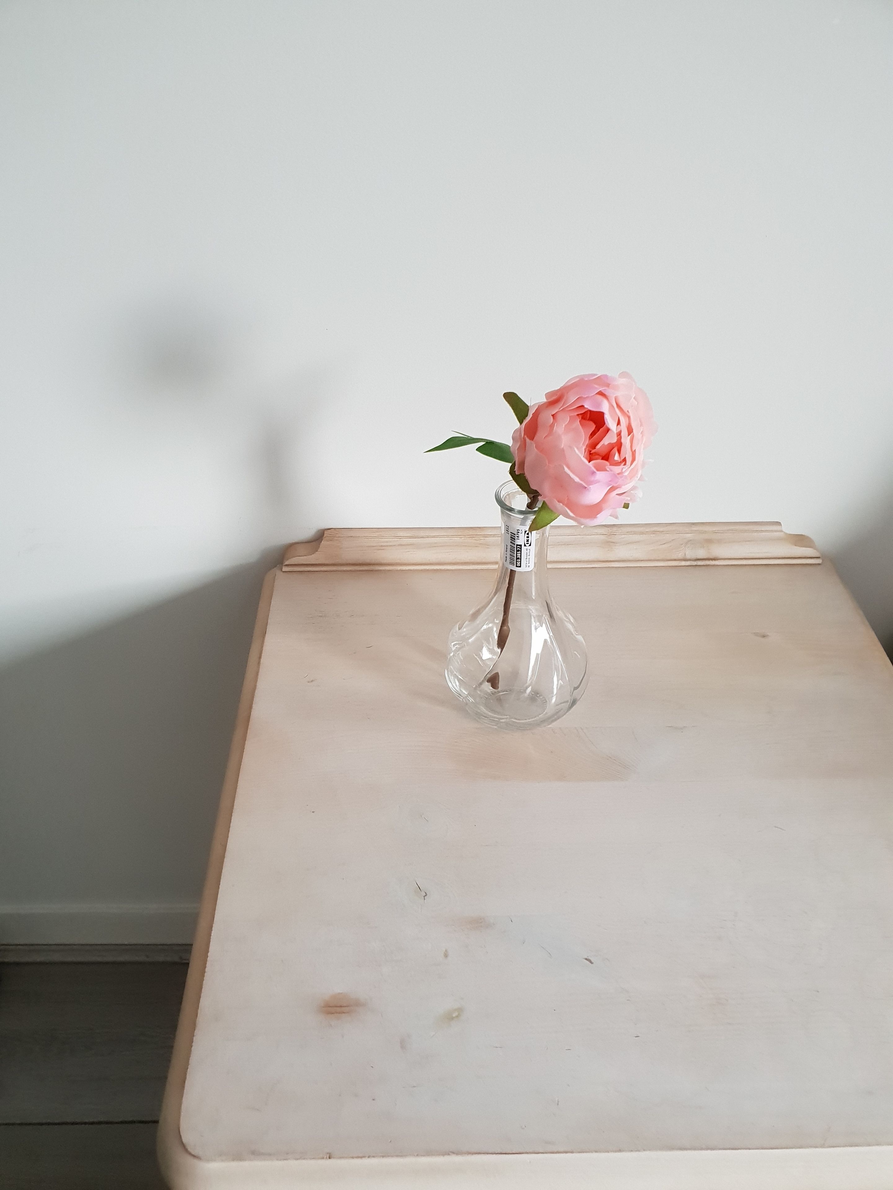 Eine transparente Glasvase mit einer rosa Blume auf einem Holztisch vor einer weißen Wand.