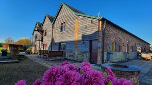 A rustic wooden building with a stone foundation, a porch with benches, and a well-maintained garden with purple flowers. - The Victorian Barn self catering holidays with pool & hot tubs (Blandford Forum)