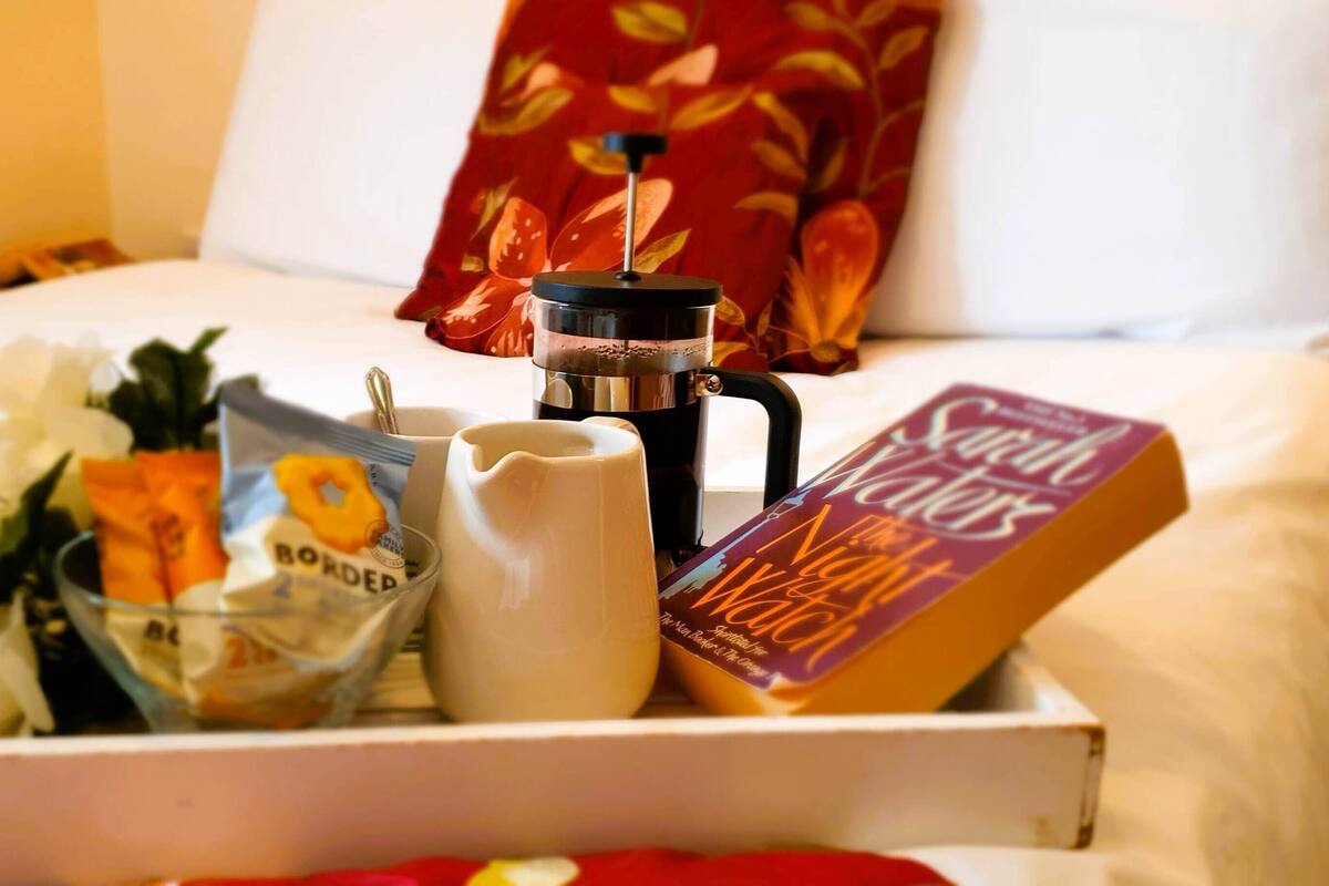 A tray with a coffee maker, a book titled 'The Night Watch', a white pitcher, and a bag of snacks on a bed.