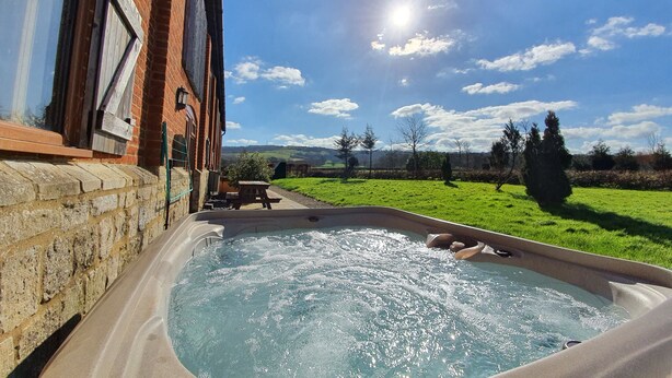 A hot tub in a backyard with a brick wall, a wooden deck, and a view of a green field and trees under a clear sky. - The Victorian Barn self catering holidays with pool & hot tubs (Blandford Forum)