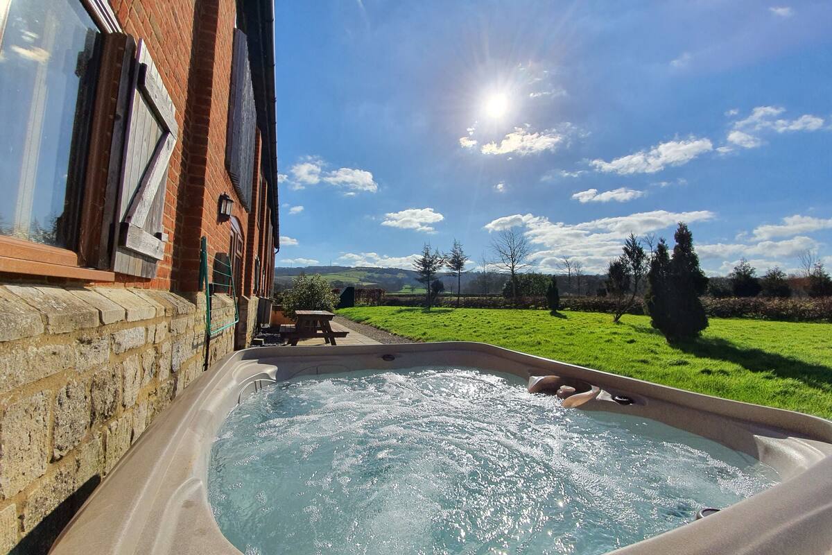 A hot tub in a backyard with a brick wall, a wooden deck, and a view of a green field and trees under a clear sky.