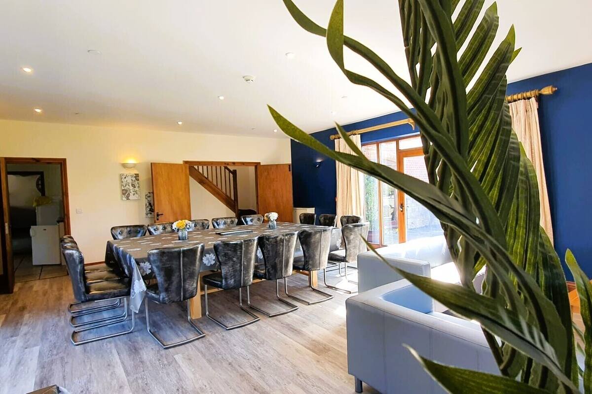 A spacious dining area with a large round table and modern chairs, a staircase leading to another level, and a large potted plant in the foreground.