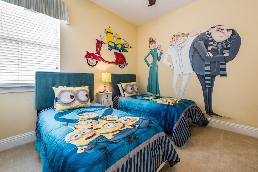 A bedroom with two beds, each with a blue bedspread featuring Minions, a wall mural of Minions and characters, and a window with blinds.