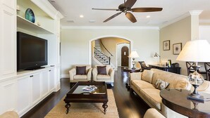 A spacious living room with a large flat-screen TV, a ceiling fan, and a staircase leading to another level.
