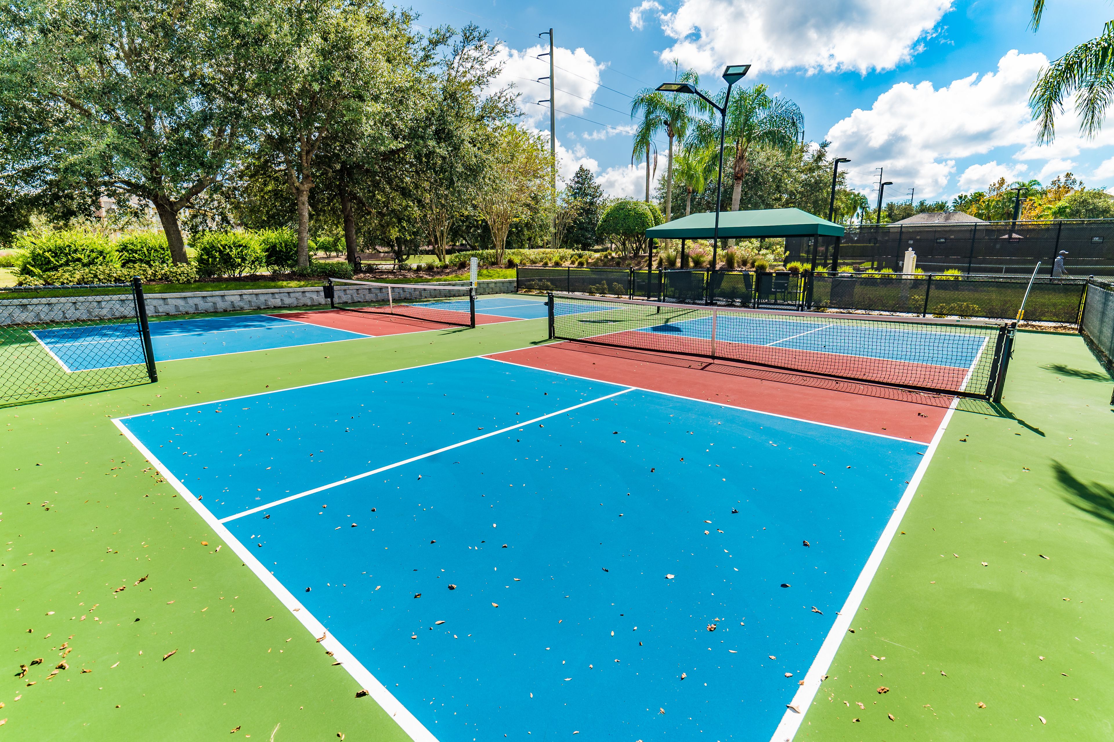 A tennis court with a green and blue surface, surrounded by trees and a covered seating area.