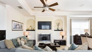 A living room with a grey sofa, a fireplace, a ceiling fan, and a television.