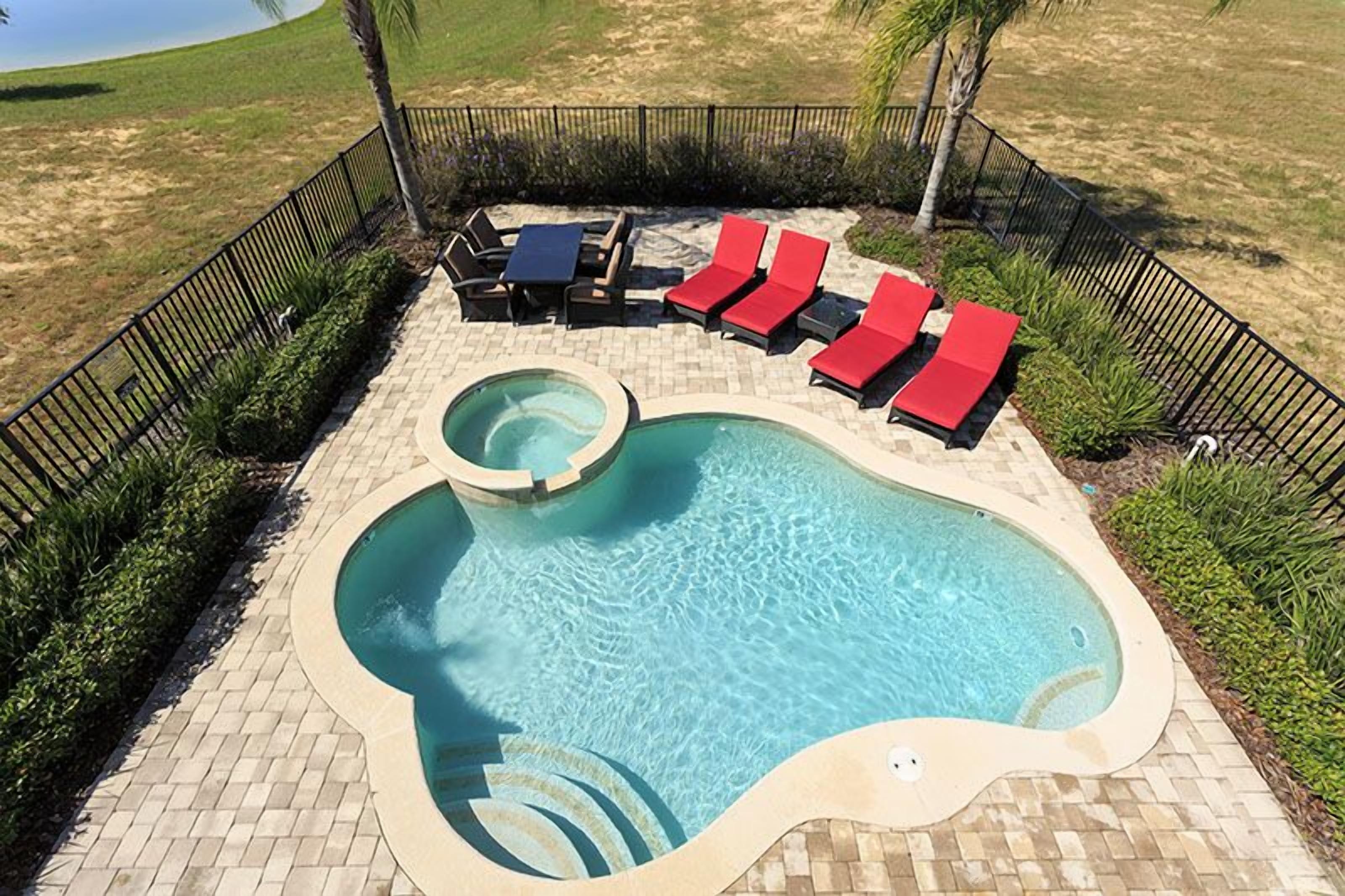 A swimming pool with red lounge chairs and a table, surrounded by a brick patio and a black fence.