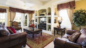 A living room with a brown leather sofa, a wooden coffee table, and a television on a stand. There is a large plant and a chandelier. - Je47858 - Reunion Resort - 6 Bed 4 Baths Villa (Kissimmee)