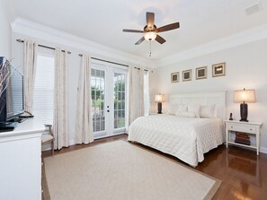 A bedroom with a bed, two bedside tables with lamps, a ceiling fan, and a large window with curtains.
